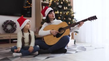 Noel Baba şapkası takan Asyalı anne ve kız Noel şarkıları söyleyerek gitar çalmanın keyfini çıkarıyorlar. Noel ağacının yanında ışık ve süslemelerle birlikte otururlar..