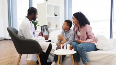 Afrikalı doktor modern klinikte endişeli anne ve oğluyla röntgen sonuçlarını tartışıyor. Anne dikkatle dinlerken erkek çocuk dijital tablete ilgi gösteriyor..