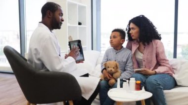 Afrikalı erkek doktor, hasta Afrikalı erkek çocuğu tablette röntgen göstererek muayene ediyor. Çocuk oyuncak ayıyı tutuyor, medikal ortamında Afrikalı yetişkin kadınların yanında oturuyor..