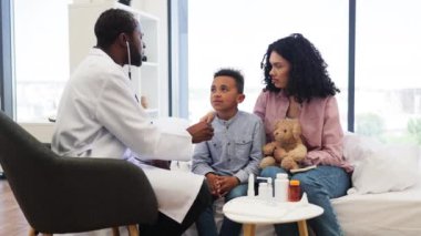 Afrikalı erkek doktor, anne yanında otururken Afrikalı genç çocukların kalp atışlarını incelemek için steteskop kullanıyor. Modern tıp bürosunda kontrol sırasında oyuncak ayıyı elinde tutan bir çocuk..
