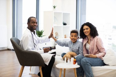 Genç Afrikalı çocuk başarılı tedaviyi kutlamak için tıbbi muayene sırasında erkek doktora beşlik çakıyor. Erkek anne, Afrikalı bir kadın, modern klinikte onun yanında gülümsüyor..