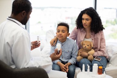 Afrikalı erkek doktor, annesinin eşlik ettiği genç Afrikalı çocuğu hastane odasında muayene ediyor. Doktor ilaç sunarken, anne elinde oyuncak ayıyla oğlunu destekliyor..