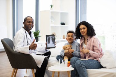 Afrikalı erkek doktor, genç Afrikalı çocuk ve beyaz anne babasıyla birlikte hastane ortamında röntgen tutuyor. Doktor, çocuk ve ebeveyn olumlu sonuç veriyor..