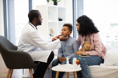 Afrikalı erkek doktor hasta Afrikalı erkek çocukların lenf nodlarını incelerken anne oyuncak ayıyla yakınlarda oturuyor. Parlak sağlık hizmetleri ortamında, tedavi ve teşhisin desteklenmesinde tıbbi danışmanlık yapılır.