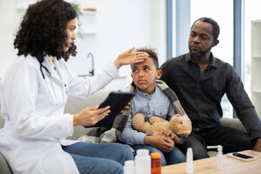 Afrikalı Amerikalı kadın doktor endişeli babasıyla oturan çocuğun ateşini ölçüyor. Çocuk, sağlık hizmetleri ve ebeveyn bakımı konseptini gösteren tıbbi bir ziyarette oyuncak ayıyı tutuyor..