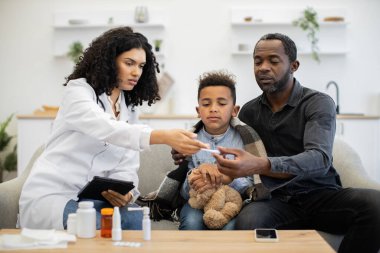 Afrikalı kadın doktor, Afrikalı babasıyla otururken çocuğun ateşini ölçüyor. Sahne, evde sağlık, aile bakımı ve konforu betimliyor.