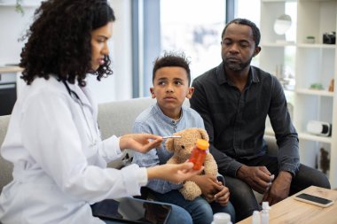 Afrikalı kadın doktor, babasıyla kanepede oturan küçük hasta çocuğa ilaç veriyor. Çocuk doktoru dinlerken oyuncak ayıyı tutuyor. Olay yeri, aile ortamında sağlık ve aile desteğini temsil ediyor