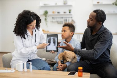 Afrikalı kadın doktor babasıyla küçük hasta çocuğa tablette akciğer röntgeni gösteriyor. Aile kanepeye oturmuş, sağlık sorunlarını tartışıyorlar. Doktor steteskop takıyor, hasta bakımıyla meşgul..