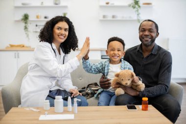 Beyaz önlüklü Afrikalı kadın doktor babalarının kanepesinde oturan küçük çocuğa beşlik çakıyor. Gülümseyen çocuk oyuncak ayıyı tutuyor, ev ziyareti sırasında neşe ve rahatlık gösteriyor..