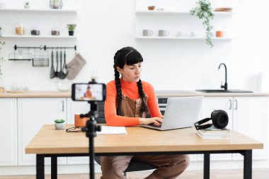 Modern mutfaktan dizüstü bilgisayarı ve akıllı telefonu olan genç bir kadın. Uzaktan çalışma ayarları kulaklıklar, telefon tripodu ve konforlu ortam içerir.