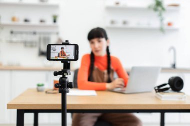 Genç bayan içerik yaratıcısı modern mutfak ortamında video kaydediyor. O, yemek masasında dizüstü bilgisayarla otururken tripodda akıllı telefon kullanıyor..