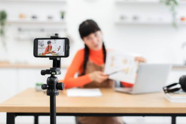 20 'li yaşlarda beyaz kadın nüfuzlu biri ilgi çekici bir içerik yaratıyor. Tripod 'a monte edilmiş akıllı telefon ve masadaki dizüstü bilgisayarı kullanıyor. Sahne yaratıcılığı ve modern dijital yaşam tarzını yansıtır.
