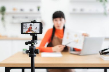 30 'lu yaşlardaki beyaz kadın nüfuzlu. Üç ayaklı akıllı telefonlu video dersleri alıyor. Masa başında oturmuş, belgeyi işaret ediyor. Parlak, modern bir ortamda geçiyor..