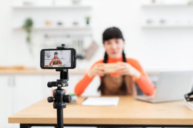 Beyaz bir kadın, mutfak ortamında yaşam tarzı blogu için boks videosu hazırlıyor. Genç kadın nüfuzlu kişi çevrimiçi izleyiciler için bağlanan içeriği yakalamak için akıllı telefonu tripoda kullanıyor.