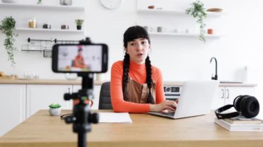 Modern mutfağın ahşap masasında dizüstü bilgisayar kullanarak oturan 20 'li yaşlarda beyaz bir kadın. Tripod üzerindeki kamera içerik oluşturma veya çevrimiçi iletişim önerir.