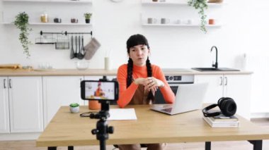 Modern mutfakta dizüstü bilgisayarla çalışan beyaz bir kadın. Gayri resmi turuncu kazak ve örgü saç stiliyle dijital içerik yaratmaya odaklanıyor. Akış için akıllı telefon ayarlandı.