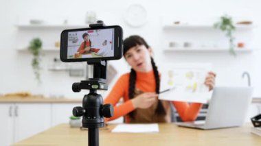 20 'li yaşlarda beyaz kadın nüfuzlu biri ilgi çekici bir içerik yaratıyor. Tripod 'a monte edilmiş akıllı telefon ve masadaki dizüstü bilgisayarı kullanıyor. Sahne yaratıcılığı ve modern dijital yaşam tarzını yansıtır.