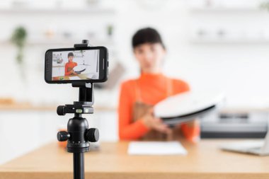 20 'li yaşlarda beyaz bir kadın parlak turuncu kıyafetli bir robot vakumunu eleştiriyor. Tripod kayıtlarındaki akıllı telefon. Modern temizlik cihazının özelliklerini tartışırken gözden geçir..