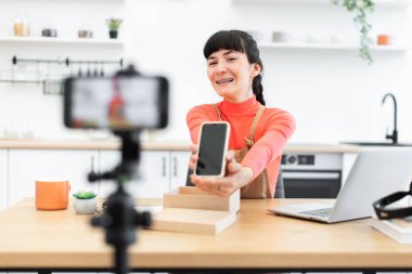 20 'li yaşların başında beyaz bir kadın en son akıllı telefon modelini internette gösteriyor. Parlak mutfaktaki ahşap masada oturmak, video kaydederken coşkuyu ifade etmek..