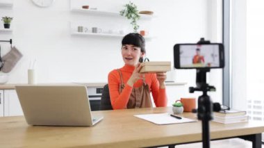 20 'li yaşların başında beyaz bir kadın en son akıllı telefon modelini internette gösteriyor. Parlak mutfaktaki ahşap masada oturmak, video kaydederken coşkuyu ifade etmek..