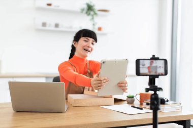 Beyaz kadın blogcu masa başında oturmuş çevrimiçi izleyicileri için yeni dijital tabletleri açıyor. Genç kadın tripoda takılı akıllı telefon kullanarak yaşıyor, dizüstü bilgisayarı, defteri ve masadaki kahveyle.