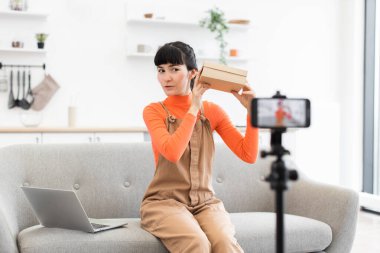 20 'li yaşlarda beyaz bir kadın video çekerken kameraya hediyesini bırakıyor. Gri kanepe ve dizüstü bilgisayarla aydınlık, modern bir iç mekanda çevrimiçi izleyicilere ilgi gösteriyor..