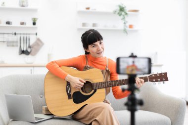 Genç Asyalı kadın çevrimiçi video kursunda gitar dersi veriyor. O, akustik gitarla kanepede oturuyor, akıllı telefonuyla içeriği kaydediyor. Parlak, modern ev tasarımı gelişigüzel gelişiyor..