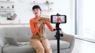 Beyaz kadın blogcu akıllı telefonuyla çevrimiçi izleyicilere gösteriyor. Modern oturma odasındaki kanepede oturmak, kamera kurulumuyla, video izlerken sevinç ifade etmek.