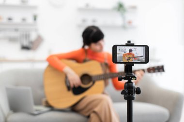 Beyaz bir kadın evde akustik gitarla video kursuna kayıt yapıyor. Video blogu için akıllı telefondan yakalanmış. Rahat oturma odası ayarları gelişigüzel öğrenmeyi ön plana çıkarır.