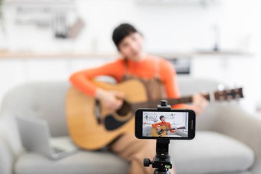 Kafkas kadın blogcu rahat ev ortamında akustik gitar çalma üzerine çevrimiçi video kursu kaydediyor. Turuncu süveter giyen kadın gitarlı koltuğa oturmuş, akıllı telefon kullanarak içerik yaratıyor..