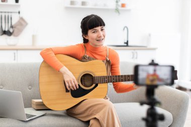 20 'li yaşlarda beyaz bir kadın evde akustik gitar çalmak üzerine çevrimiçi video kursu alıyor. Kanepede gitar, dizüstü bilgisayar ve akıllı telefon kamerasıyla oturuyor..