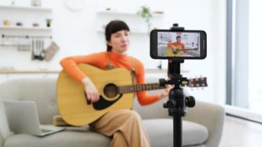 Beyaz kadın blogcu evde akustik gitar çalma üzerine çevrimiçi video kursu kaydediyor. Genç bir kadın elinde bir gitar tutarak koltukta oturuyor. Üç ayaklı telefondan talimat içeriğini yakalıyor..