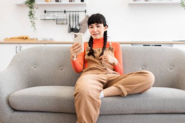 Kanepede oturan beyaz bir kadın internetten video görüşmesi yapıyor. O, turuncu bluzlu ve tulumlu günlük kıyafetler giyer. Parlak ve modern oturma odası..