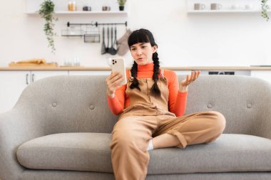 Kafkasyalı genç bir kadın rahat bir koltukta oturmuş akıllı telefon kullanarak online video görüşmesi yapıyor. Rahat ev ortamı, parlak, rahat kıyafetler..