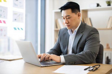 Modern ofis ortamında dizüstü bilgisayarla çalışan Asyalı iş adamı. Bilgisayar görevlerine odaklanmış takım elbiseli genç bir profesyonel. Kitaplık ve dokümanların olduğu ofis ortamı.