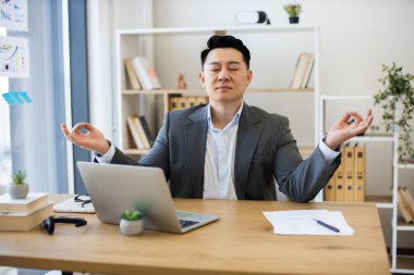 Asyalı yetişkin erkek, stres atmak için masada meditasyon yapıyor. Gözleri kapalı bir takım elbise giymek, rahatlama teknikleri uygulamak. İş ortamında sükunet ve odaklanma kavramları.