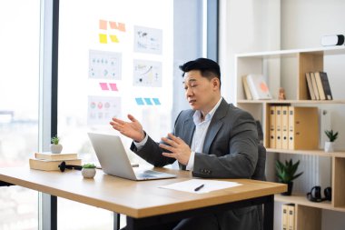 Takım elbiseli Asyalı erkek işadamı, ofiste dizüstü bilgisayar kullanıyor. Masada oturmuş, online video görüşmeleri yapıyor, iş konularını tartışıyoruz. Şehir manzaralı parlak modern ofis.