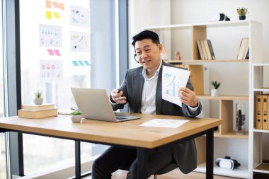 Çevrimiçi iş görüşmesine katılan Asyalı profesyonel erkek kulaklık takıyor. Ofis masasında dizüstü bilgisayarla oturuyor, elinde grafikler ve çizelgelerle iş raporları var..