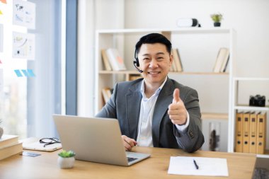 İnternetteki video görüşmesi için bilgisayarını kullanan Asyalı bir iş adamı. Kulaklık takıp, resmi takım elbise giyip kameraya baş parmak işareti yapıyor. Ofis kurulumu kitaplı, pencereli ve masa başlıklı rafları içerir.
