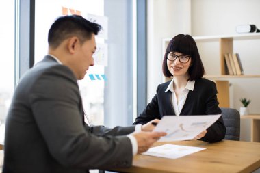 Asyalı işadamı, modern ofis ortamında beyaz iş kadınına belge sunuyor. İkisi de resmi iş kıyafeti giyiyor, profesyonel bir tartışma içindeler..