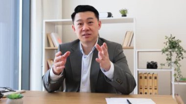 Modern ofisteki Asyalı işadamı online video görüşmesine dizüstü bilgisayarla bağlandı. Profesyonel ayarlar arasında duvardaki dijital çizelgeler, ince ışıklandırma ve düzenli masa kurulumu yer alıyor.