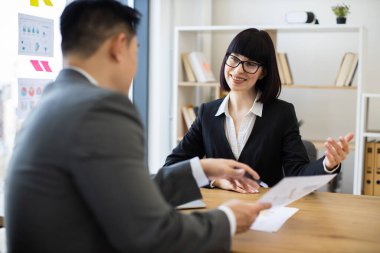 Asyalı işadamı belgeyi modern ofis ortamında beyaz iş kadınıyla paylaşıyor. Profesyonel etkileşimde takım çalışması, işbirliği ve iletişim vurgulanır.