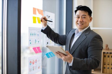 Asyalı erkek işadamı tablet kullanarak panoramik pencere üzerindeki grafikleri ve grafikleri analiz ediyor. Orta yaşlı profesyonel takım elbise giyiyor, renkli yapışkan notlar ve belgelerle çalışırken gülümsüyor..