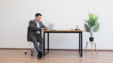 Resmi takım elbiseli Asyalı iş adamı ahşap masada otururken dizüstü bilgisayarda çalışıyor. Minimalist dekorlu modern ofis ortamında saksı bitkileri ve şık mobilyalar da dahil..