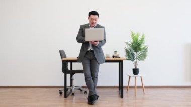 Asyalı erkek işadamı dizüstü bilgisayar kullanarak ofiste güven içinde duruyor. Şık bir iş kıyafeti giymiş, nişanlı ve gülümsüyor..