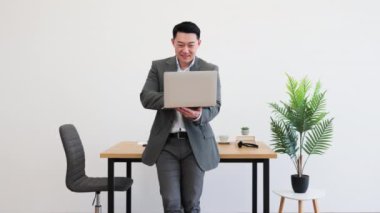 Asyalı profesyonel erkek, minimalist ofiste dizüstü bilgisayar tutuyor ve gülümsüyor. Meşgul ve kendinden emin görünüyor, iş kıyafeti giymiş..