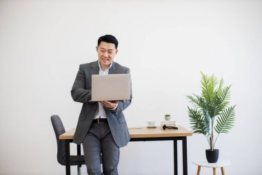 Asyalı profesyonel erkek, minimalist ofiste dizüstü bilgisayar tutuyor ve gülümsüyor. Meşgul ve kendinden emin görünüyor, iş kıyafeti giymiş..