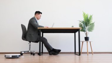 Takım elbiseli Asyalı erkek işadamı beyaz duvar arkasında masada otururken dizüstü bilgisayarda çalışıyor. Modern ofis dekorasyonunda ahşap zemin ve yeşil fabrika.