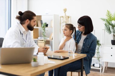 Tıbbi konsültasyon sırasında annesinin yanında oturan endişeli yüzlü beyaz bir kız. Bayan ebeveyn ve kızı, modern, parlak tıp ofisinde erkek doktorla tartışıyorlar..