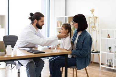 Erkek doktor modern klinikte annesiyle masada oturan beyaz bir kızın lenf nodlarını inceliyor. Kadın gözlük takıyor, endişeleniyor. Odada tıbbi çizelge ve iskelet var..